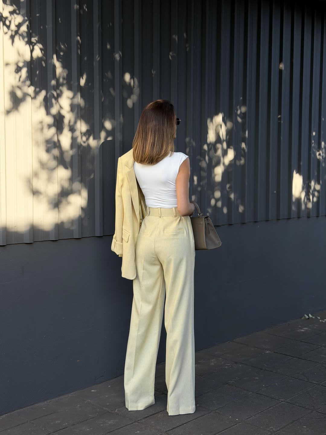 Look de alfaiataria amarelo-manteiga visto de costas, com blazer sobre o ombro e bolsa bege — ZAYA Boutique, Curitiba